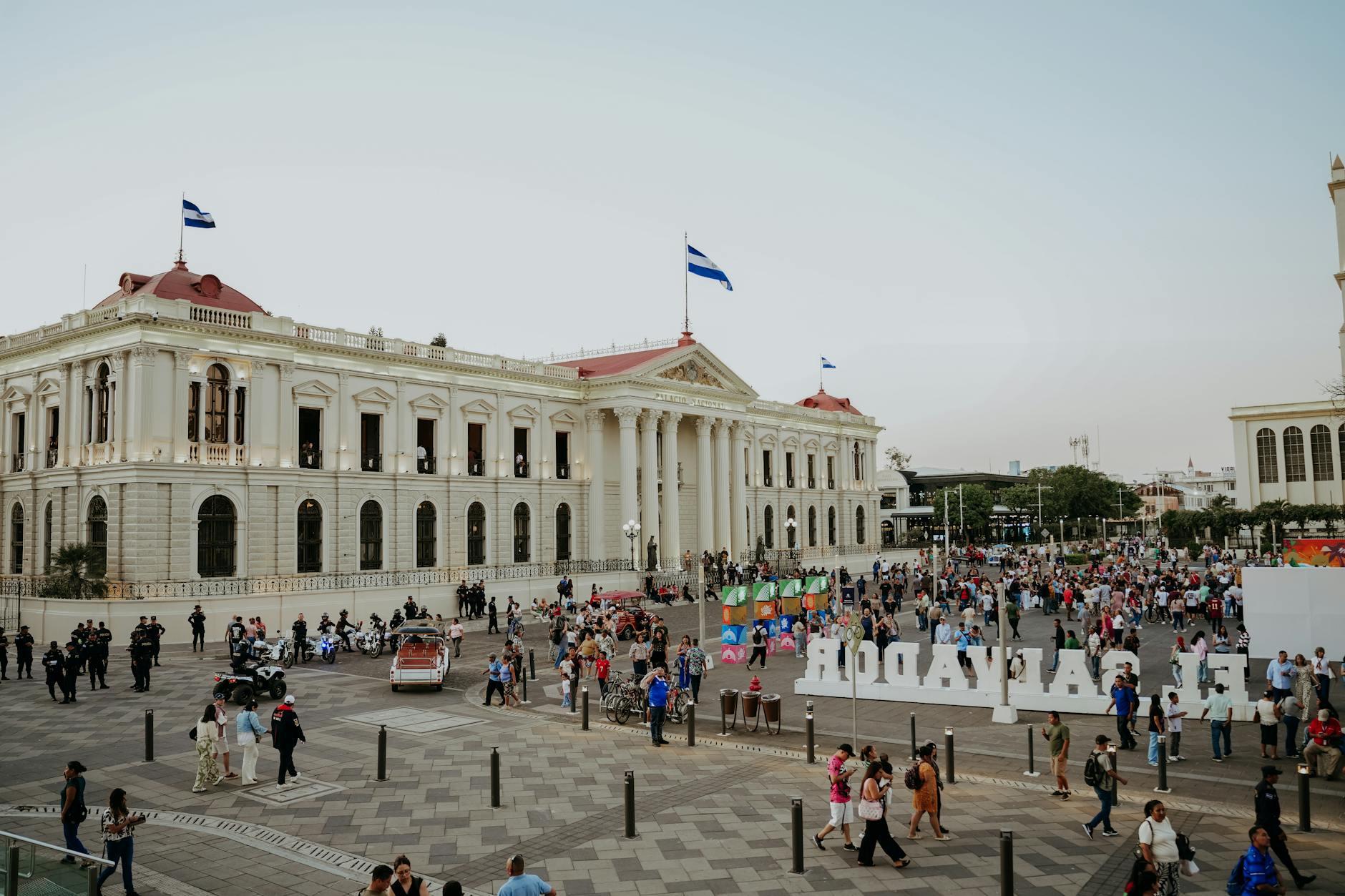 Crowded plaza scene in San Salvador with flags and historic buildings under a clear sky.